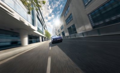 The all-new Hyundai KONA Electric in blue driving down a city street in the sun.