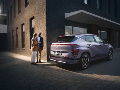 Ein vor einem Gebäude parkender Hyundai KONA Elektro in der Heckansicht – daneben ein sich unterhaltendes Paar.