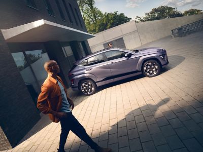 The Hyundai KONA Electric parked in the sunlight with a man walking away from a building.