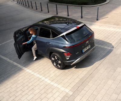 A woman crosses the street with a green Hyundai KONA is parked on the other side. 