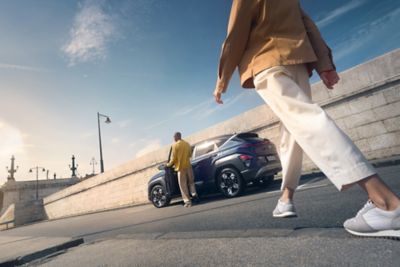 A man with a yellow shirt enters the all-new Hyundai Kona with a person walking towards to it.	