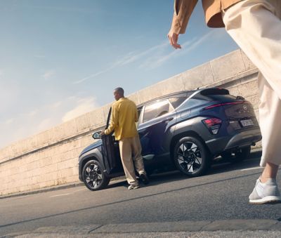 A man with a yellow shirt enters the all-new Hyundai Kona in blue and a person walks towards to it.