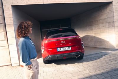 A woman with a blue shirt stands behind the all-new Hyundai KONA in red parked inside a garage. 