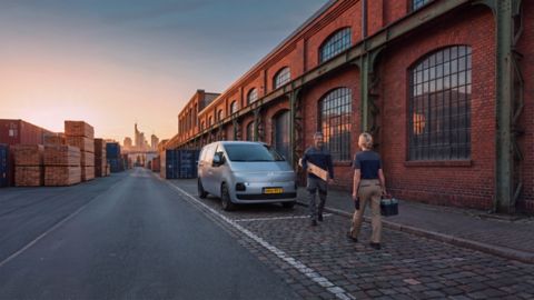 Driekwart vooraanzicht Hyundai STARIA Cargo met Nederlands kenteken geparkeerd in een industriële omgeving met op de voorgrond een man en vrouw in werkkleding die op elkaar aflopen.