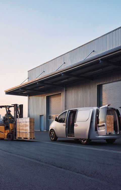 En STARIA Electric Varebil står parkert ved er lager og en truck kjører forbi. Foto.