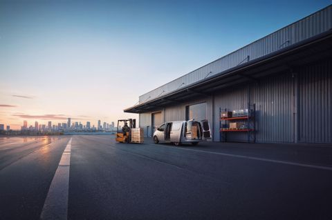 En STARIA Electric Varebil står parkert ved er lager og en truck kjører forbi. Foto.