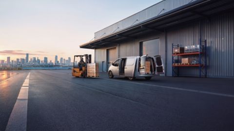 Driekwart achteraanzicht Hyundai STARIA Cargo met geopende schuifdeur en geopende dubbele achterdeuren, geparkeerd in een industriële omgeving met een vorkheftruck die komt aanrijden.