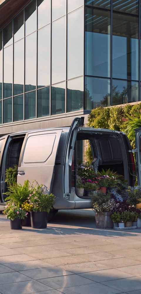 En kvinne losser potteplanter fra elektriske Hyundai STARIA Cargo utenfor en moderne bygning.