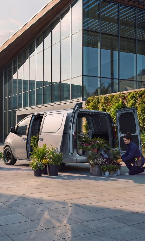 En kvinne som laster ut potteplanter fra en Hyundai STARIA Electric Varebil utenfor en moderne bygning.
