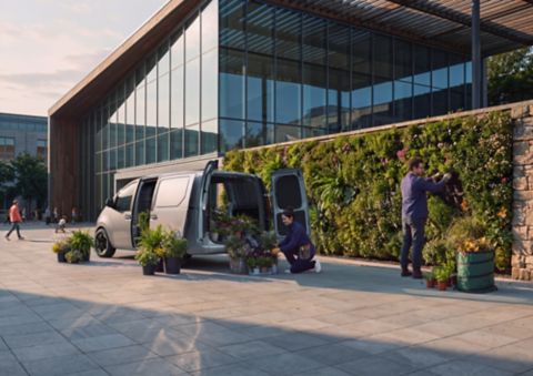 En kvinne laster ut potteplanter fra elektriske Hyundai STARIA Electric Varebil utenfor en moderne bygning.