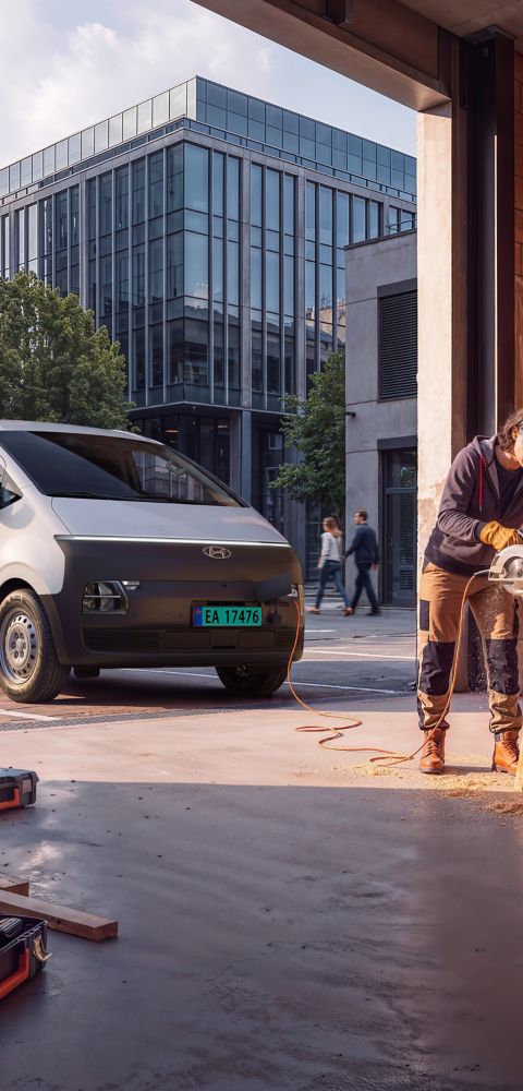 Hyundai STARIA Electric Varebil parkert ved et verksted mens en arbeider kapper trevirke på en arbeidsplass i byen.