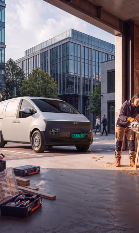 Hyundai STARIA Electric Varebil står parkert ved et verksted mens en arbeider kapper trevirke på en arbeidsplass i byen.