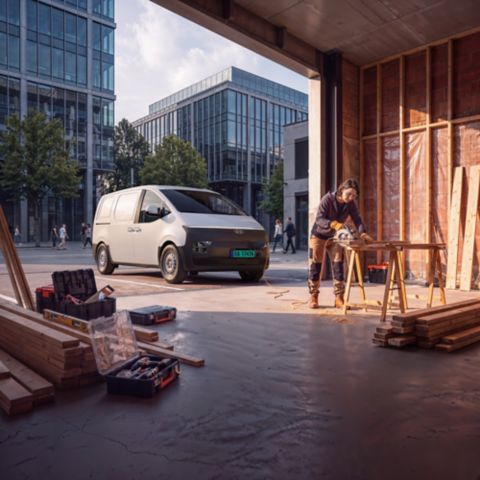 Hyundai STARIA Electric Varebil parkert ved et verksted mens en arbeider kapper trevirke på en arbeidsplass i byen.