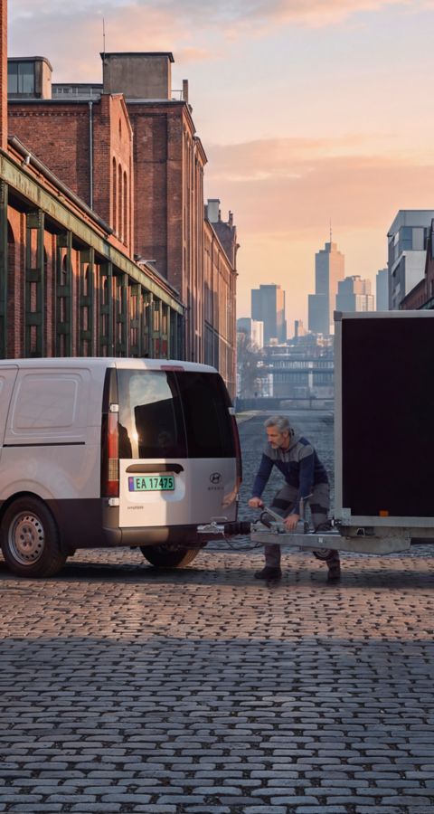 Hyundai STARIA Electric Varebil som trekker en varehenger på en brosteinsgate i et urbant industriområde.
