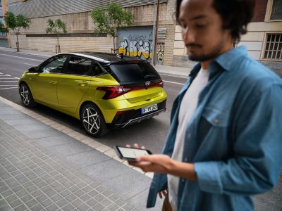 Muž používá smartphone vedle žlutého vozu Hyundai i20 zaparkovaného na ulici. 