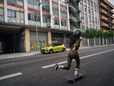Une jeune femme fait du skateboard dans la rue avec en arrière-plan, nouvelle Hyundai i20.