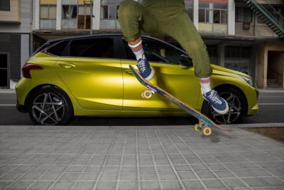 Ein Mensch mit einem Skateboard macht einen Sprung vor einem Hyundai i20 in der aufpreispflichtigen Farbe Lucid Lime Metallic.	