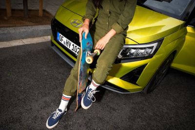 Ein Mensch mit einem Skateboard zwischen den Beinen sitzt auf der Motorhaube des Hyundai i20 in der aufpreispflichtigen Farbe Lucid Lime Metallic.