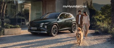 Familj promenerar med hund bredvid en Hyundai SUV med takbox framför ett hus i sommargrön miljö.