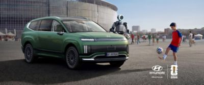 Hyundai SANTA FE parkerad utanför en fotbollsstadion tillsammans med Boston Dynamics-roboten Atlas och ett barn som spelar fotboll under FIFA World Cup 2026™-kampanjen.