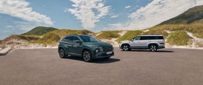 Hyundai TUCSON Plug-in Hybrid och Hyundai SANTA FE Plug-in Hybrid parkerade på en öppen asfaltsyta framför sanddyner under en klar himmel. TUCSON syns i förgrunden i en mörk färg medan SANTA FE står i bakgrunden i profil. 