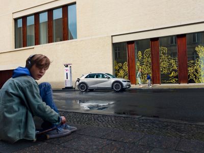 The Hyundai IONIQ 5 electric vehicle in charge and a boy sitting down the street.