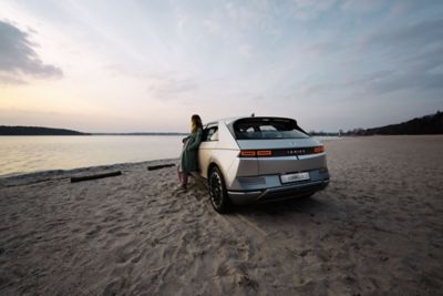 The Hyundai IONIQ 5 electric midsize CUV parked at a beach with a woman leaning against it, enjoying the sundown.