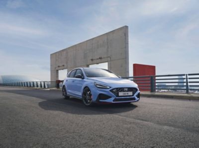 The Hyundai i30 N from the front right in Performance Blue parked next to an industrial structure.
