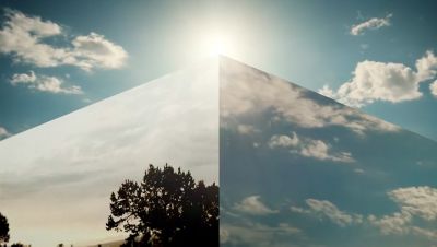 A cornered mirrored monolith reflecting a blue and cloudy sky with a tree.