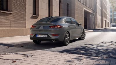 Nowy Hyundai i30 Fastback w kolorze Shadow Gray, zaparkowany w mieście – ujęcie 3/4 z tyłu.