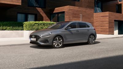 Tříčtvrteční pohled zepředu na nový Hyundai i30 v šedé barvě, zaparkovaný před moderním domem.