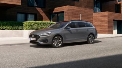 Driekwart vooraanzicht van de nieuwe Hyundai i30 Wagon in grijs, geparkeerd voor een modern huis.