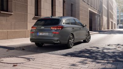 Driekwart achteraanzicht van de nieuwe Hyundai i30 Wagon in grijs, geparkeerd in een stadsstraat.