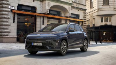 Ein blauer Hyundai KONA Hybrid, von vorne, auf einer sonnenbeschienenen Büro- und Geschäftsstraße.
