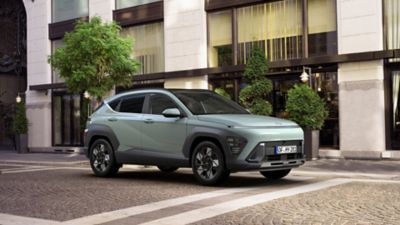 A woman crosses the street with a green Hyundai KONA is parked on the other side. 