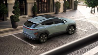 A woman crosses the street with a green Hyundai KONA is parked on the other side. 