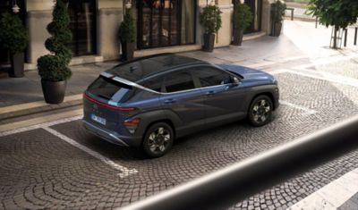 A woman crosses the street with a green Hyundai KONA is parked on the other side. 