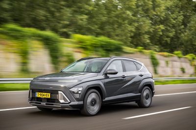 Driekwart vooraanzicht rijdende Hyundai KONA Electric op snelweg langs een geluidswal met begroeiing en bomen op de achtergrond.