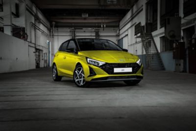 Nouvelle Hyundai i20 en jaune, de profil, stationnée dans un décor urbain moderne.