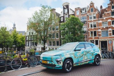 Hyundai KONA Electric gehuld in kleuren van schilderij ‘Amandelbloesem’ van Vincent van Gogh op brug over Amsterdamse gracht.