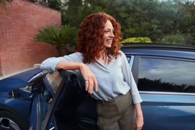 Vrouw leunt met arm op bovenzijde geopende bestuurdersportier van een geparkeerde Hyundai BAYON.