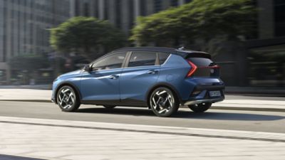 Side view of a blue Hyundai BAYON driving down a city street with trees in the background.