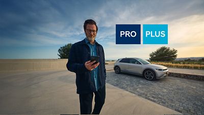 A person holding a smartphone stands near a Hyundai IONIQ 5 parked in the background. The image includes two blue labels with the words “PRO” and “PLUS,” highlighting Hyundai’s Bluelink packages.