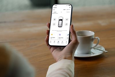 Ekranında elektrikli otomobil kontrol uygulamasının uzaktan klima kontrolü ve otomobil durumu sayfalarının görüldüğü bir akıllı telefon tutan bir el.