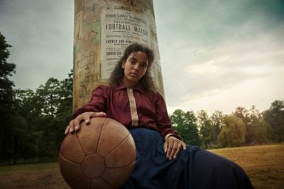 Eine Fußballspielerin sitzt mit Ball an einer Säule mit Plakat - im Stil der 20er Jahre.