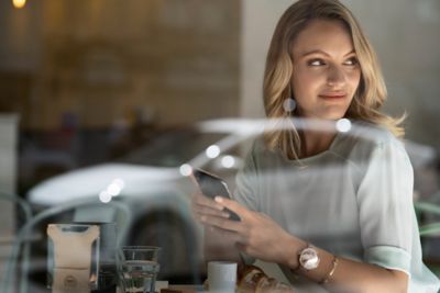 A woman looking at her Hyundai through a coffee shop window, holding her phone in her hands.