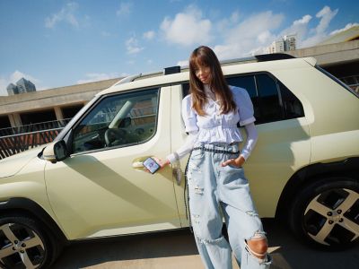 A woman leans against the new Hyundai INSTER small electric car, using Bluelink on her smartphone.