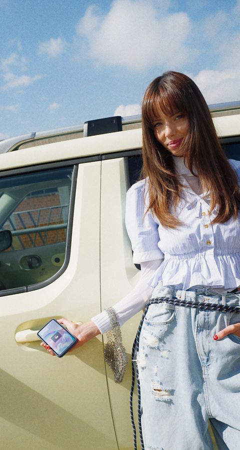 A woman leans against the new Hyundai INSTER small electric car, using Bluelink on her smartphone.