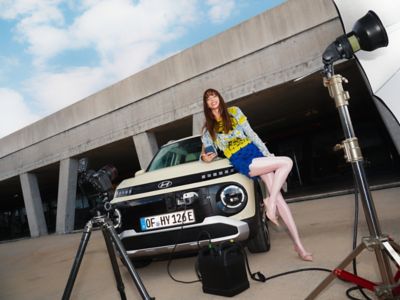 A woman poses on the new Hyundai INSTER small electric car, used as a charging station for camera equipment nearby.