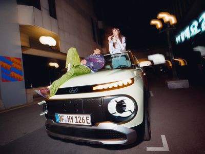 Woman standing in the new Hyundai INSTER, with a man lying on the bonnet.